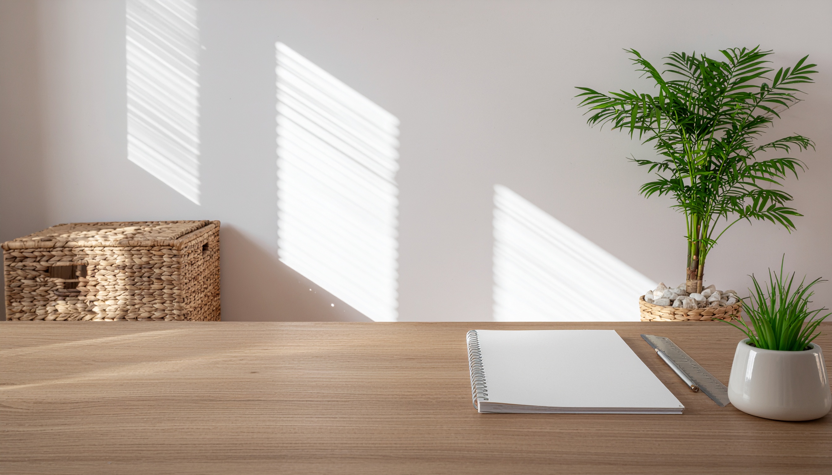Mesa de madeira clara em ambiente minimalista com caderno em espiral, régua metálica e caneta prateada. Plantas em vasos decorativos e cesta de vime ao fundo, com luz suave entrando por persianas, criando sombras geométricas na parede branca.